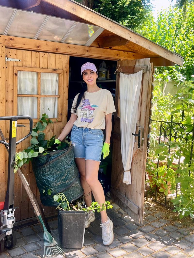 Somayeh en casquette et gants de jardinage devant un cabanon en bois, un sac de feuilles à côté d’elle